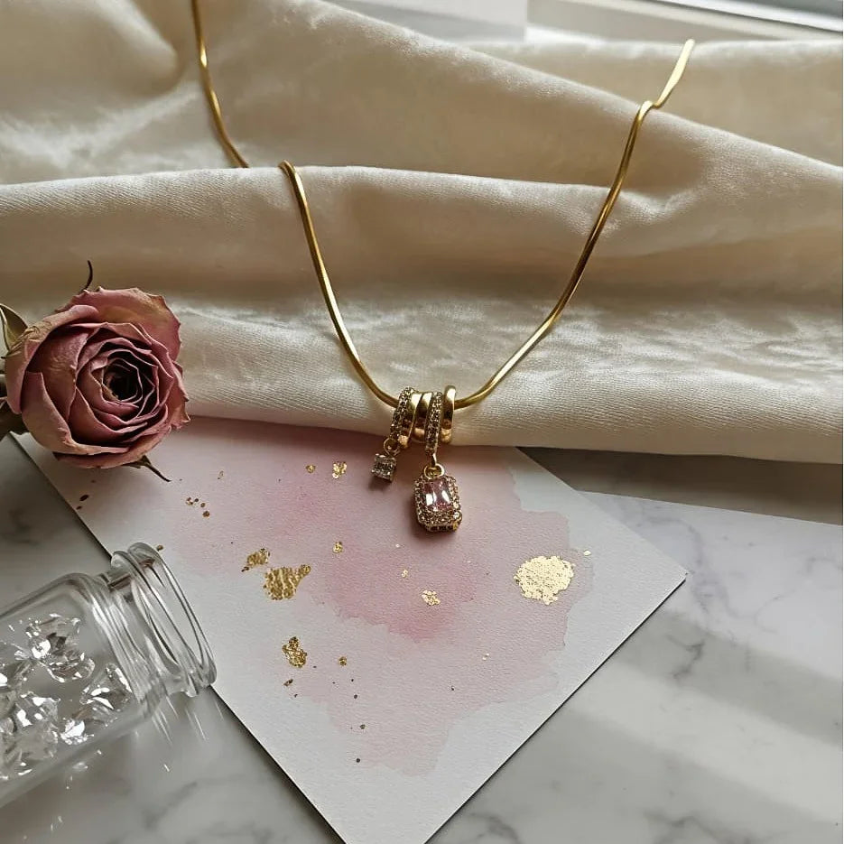 Gold necklace with pink charms on a marble surface with a pink rose and card.